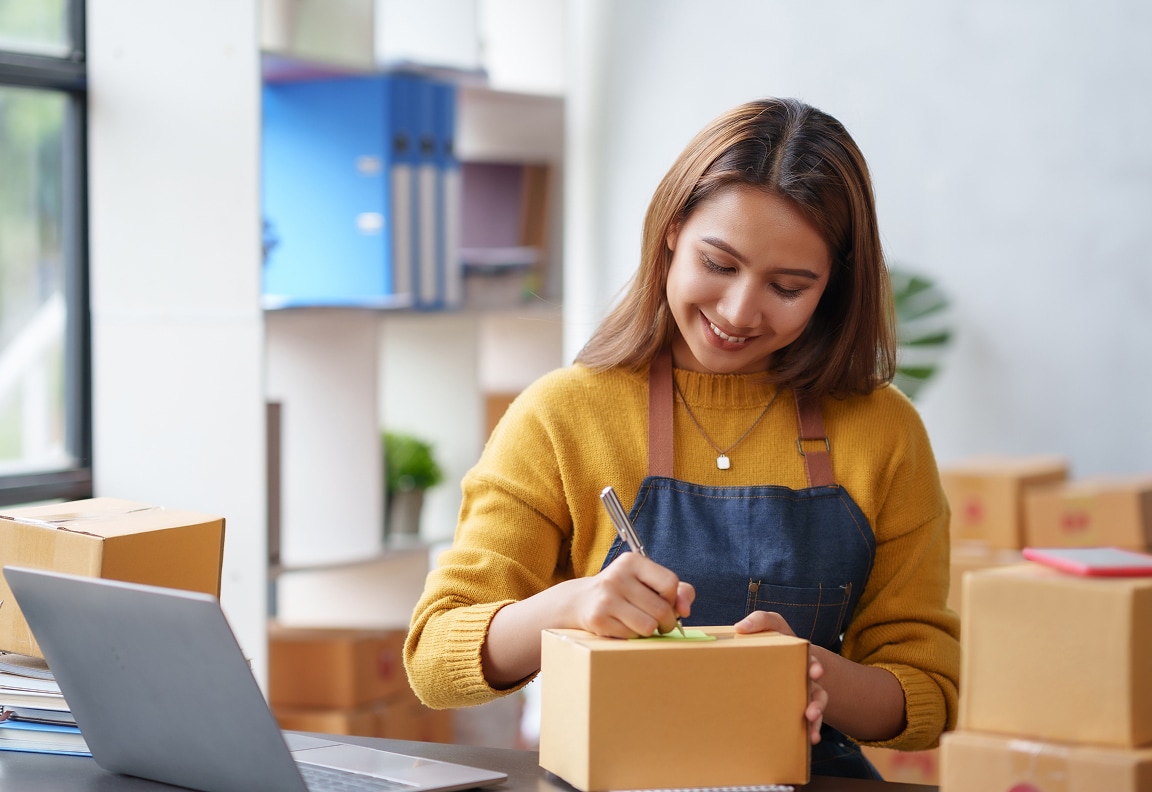 Une femme souriante est assise à un bureau et écrit sur une petite boîte. D’autres boîtes d’expédition se trouvent alentour.