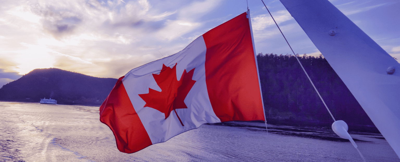 Un drapeau canadien flottant au vent, avec un plan d’eau et des collines boisées à l’arrière-plan.