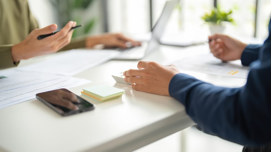 Deux personnes travaillent à un bureau. L’une utilise une calculatrice et l’autre tient un stylo en utilisant un ordinateur.