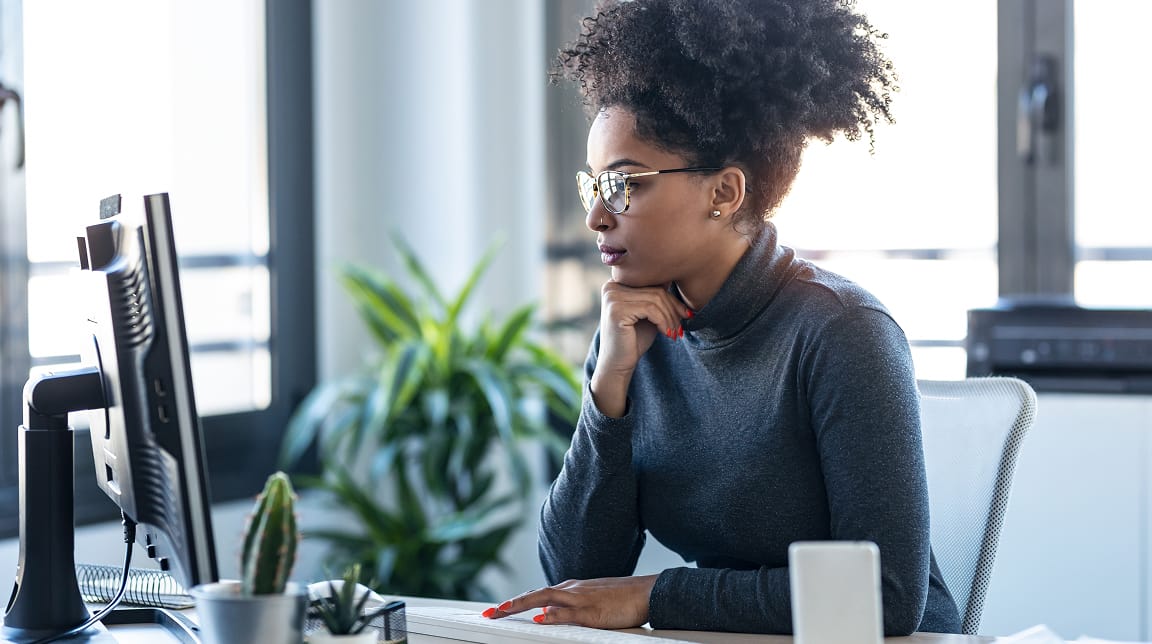 Une femme assise à un bureau regarde l’écran de l’ordinateur en reposant son menton sur son poing.