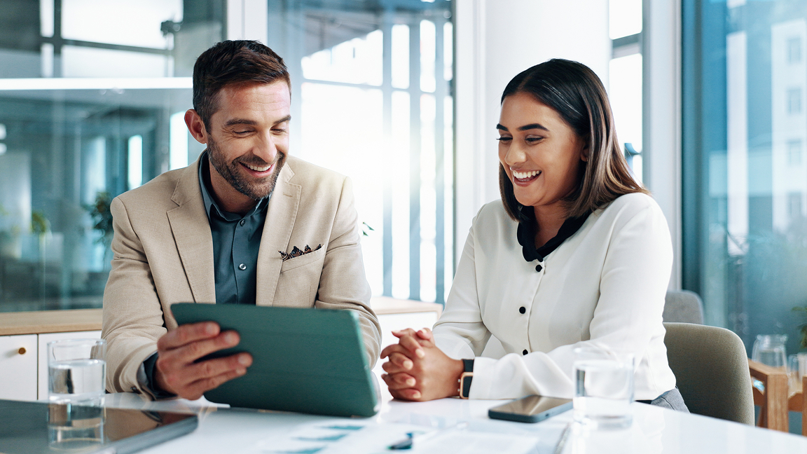 Un homme et une femme sont assis côte à côte à un bureau. L’homme tient une tablette et montre l’écran à la femme.