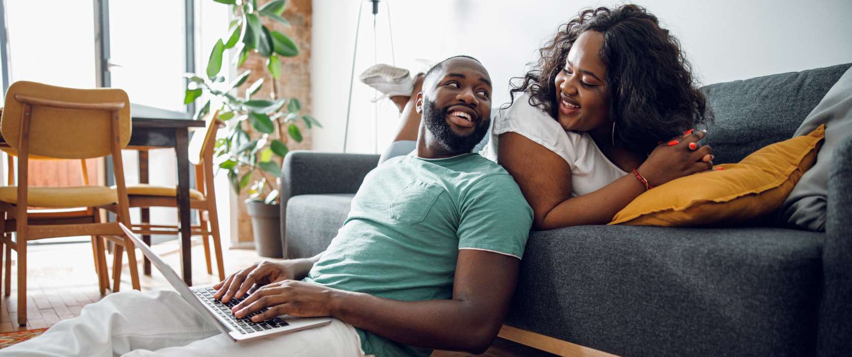 Un homme tape sur un ordinateur portable et s’appuie sur un canapé sur lequel une femme est assise. Les deux se sourient.