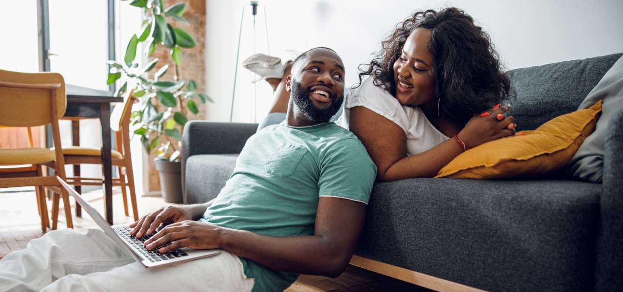 Un homme tape sur un ordinateur portable et s’appuie sur un canapé sur lequel une femme est assise. Les deux se sourient.