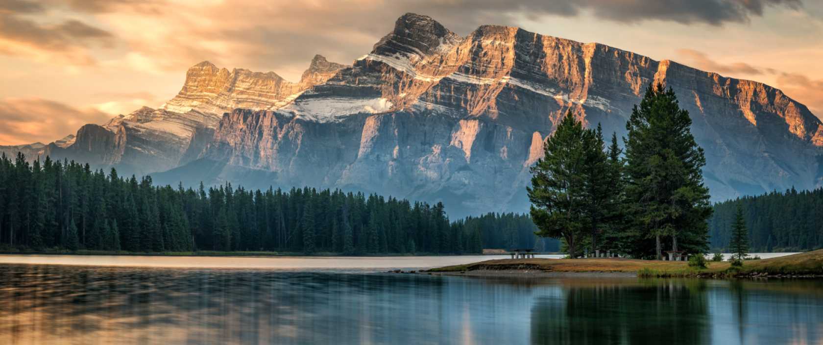 Un lac paisible entouré d’une forêt qui se situe au pied d’une montagne rocheuse et qui est baigné dans la lumière dorée d’un coucher de soleil.