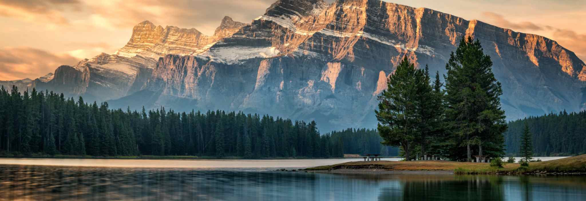 Un lac paisible entouré d’une forêt qui se situe au pied d’une montagne rocheuse et qui est baigné dans la lumière dorée d’un coucher de soleil.