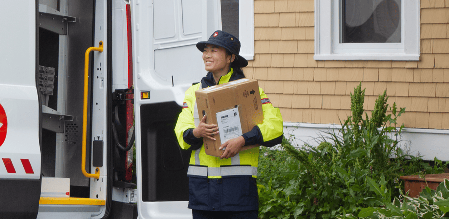 Un employé des postes en uniforme haute visibilité transporte un grand colis près d’un véhicule de livraison ouvert devant une maison.