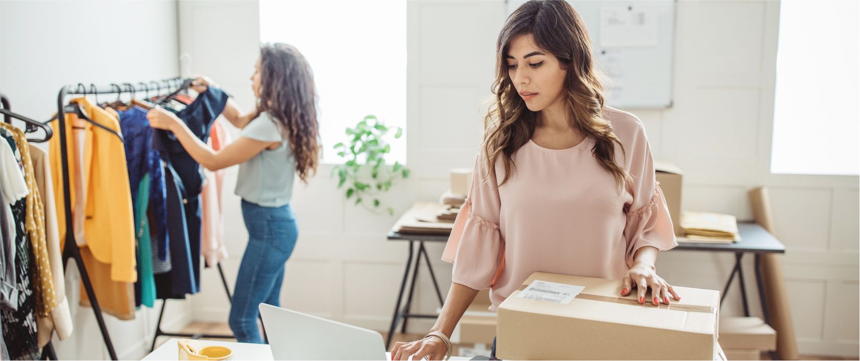 Une jeune femme laisse reposer sa main sur une boîte d’expédition. Une autre femme ajoute des vêtements à un présentoir derrière elle.