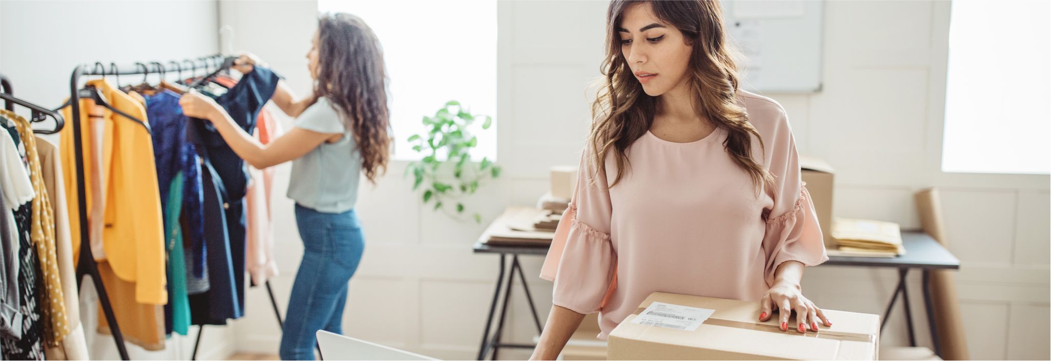Une jeune femme laisse reposer sa main sur une boîte d’expédition. Une autre femme ajoute des vêtements à un présentoir derrière elle.